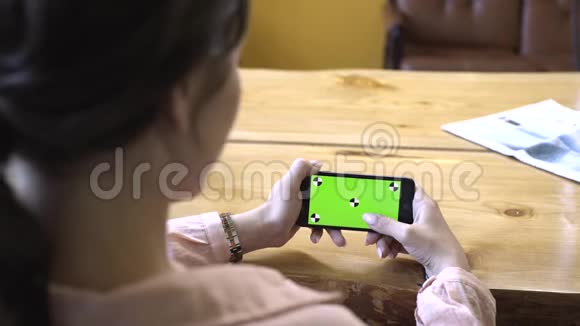 从棕色头发穿着粉红色衬衫坐在木桌旁手拿着手机的年轻女子的背影看视频的预览图