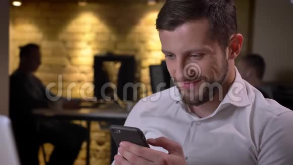 特写年轻开朗的白种人商人在手机上浏览微笑地坐在笔记本电脑前视频的预览图