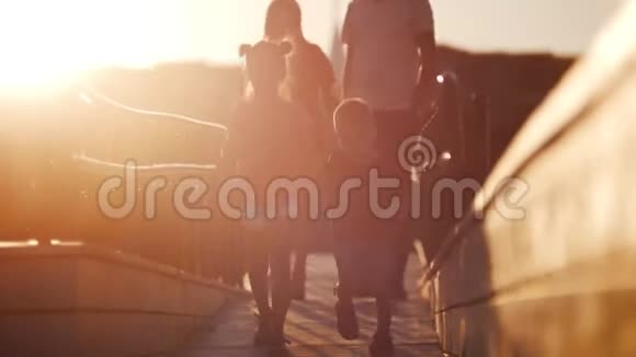 年轻的一家人靠着美丽的夕阳走在桥上孩子们跑在前面后面的父母视频的预览图