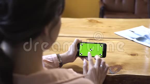 从棕色头发穿着粉红色衬衫坐在木桌旁手拿着手机的年轻女子的背影看视频的预览图