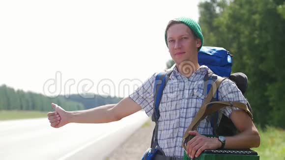 一个穿格子衬衫的年轻人一顶绿帽子和一个背包站在路边按喇叭视频的预览图