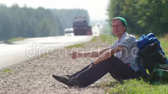 一个穿格子衬衫的年轻人一顶绿帽子和一个背包坐在路边按喇叭视频的预览图