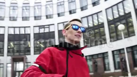 穿着红色运动服和太阳镜的帅哥在灰色的大建筑旁边跳舞视频的预览图