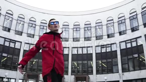 穿着红色运动服和太阳镜的年轻男性在灰色的大建筑旁边跳舞视频的预览图