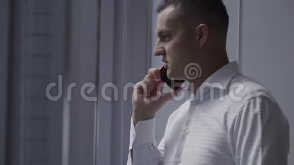 穿着白色衬衫的漂亮黑发男子在办公室的窗户附近的智能手机上讲话视频的预览图