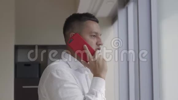 穿着白色衬衫的漂亮男人在办公室的窗户附近的智能手机上讲话视频的预览图