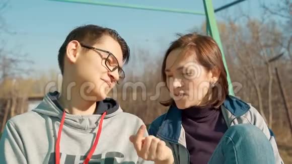 儿子和他母亲坐在露天的长凳上带着儿子的女人正在看智能手机视频的预览图
