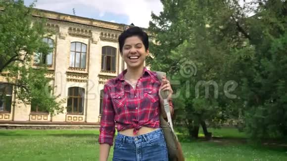 快乐的年轻女子留着短发微笑着对着镜头笑站在学院大楼附近的公园里视频的预览图