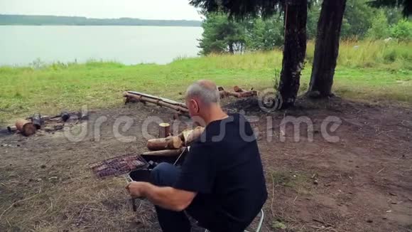 河岸上的人坐在烤肉旁翻着煤手里拿着烤架和鱼视频的预览图