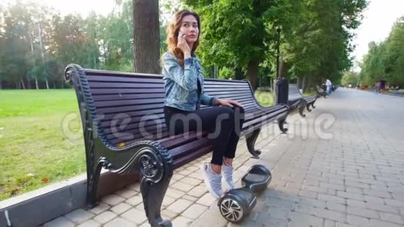 年轻苗条的女孩坐在长凳上打电话然后她坐着陀螺从长凳上站起来骑车视频的预览图