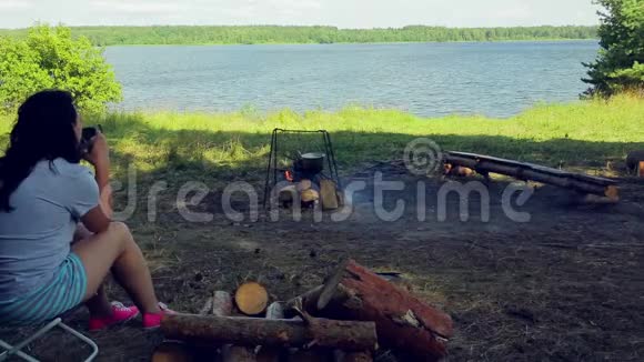 一个女人坐在火边的河岸上喝茶视频的预览图