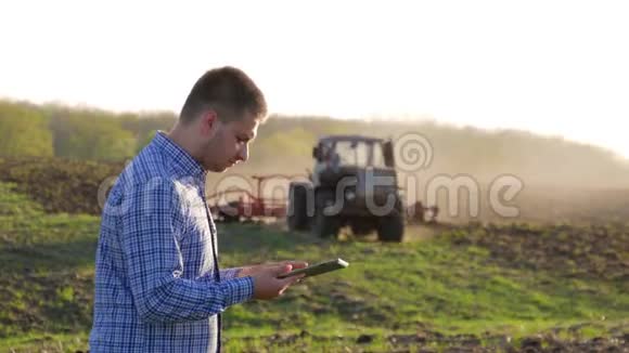 年轻的农民在田间拖拉机前拿着平板电脑农夫用平板电脑控制着田里工作视频的预览图