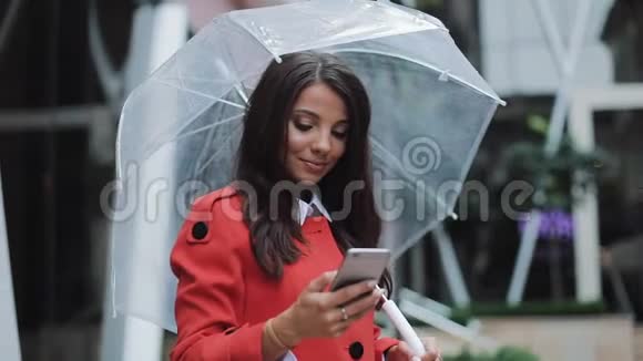 快乐的年轻商务女士站在城市街道上穿着红色外套使用智能手机她拿着雨伞视频的预览图