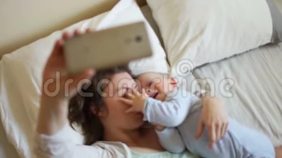 幸福的妈妈和宝宝躺在床上拍摄摄像机手机视频的预览图