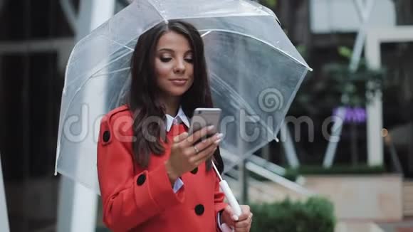 快乐的年轻商务女士站在城市街道上穿着红色外套使用智能手机她拿着雨伞视频的预览图