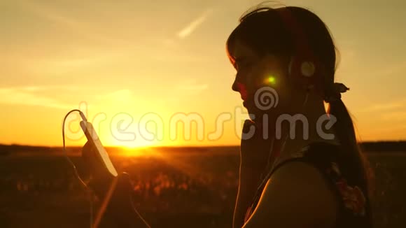 快乐的女孩在智能手机上听音乐在美丽的春天日落的光线下慢动作年轻女孩视频的预览图