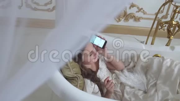 美丽的年轻女子躺在安瓿浴缸的枕头上用手机聊天视频的预览图