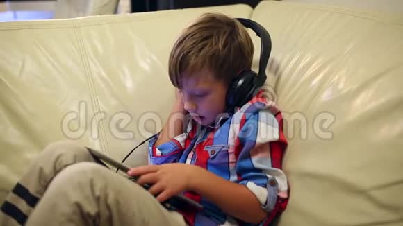 快乐的小男孩戴着耳机在平板电脑上播放音乐坐在家里的沙发上听音乐视频的预览图