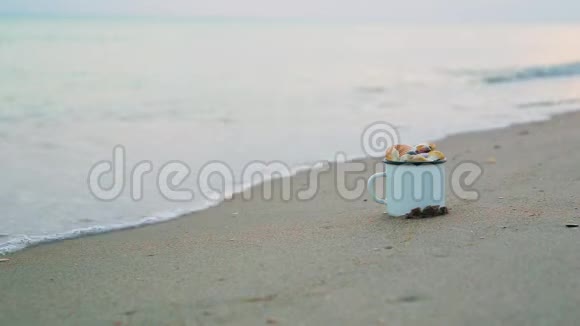 一个白色的金属杯里面装满了贝壳矗立在沙质的海岸上面对着模糊的海浪视频的预览图