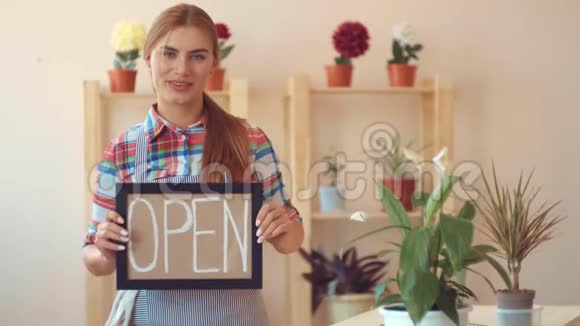 小生意开一家花店穿格子衬衫和条纹围裙的女孩有一个开放的标志在后台视频的预览图