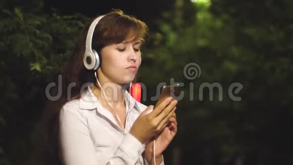 戴着耳机的漂亮女孩在智能手机上听音乐和跳舞1女孩晚上在城市公园散步视频的预览图