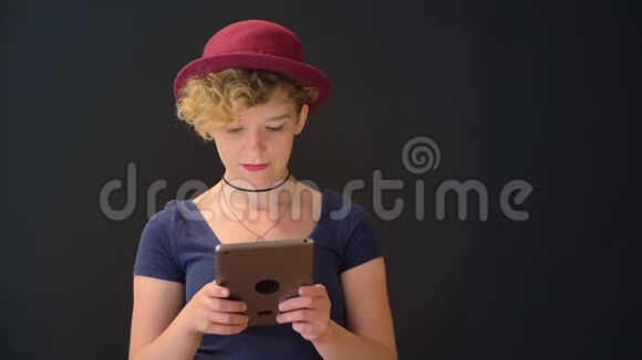 戴着红帽子卷发拿着平板电脑看着摄像机的严肃年轻美丽的女人被隔离在黑色上视频的预览图