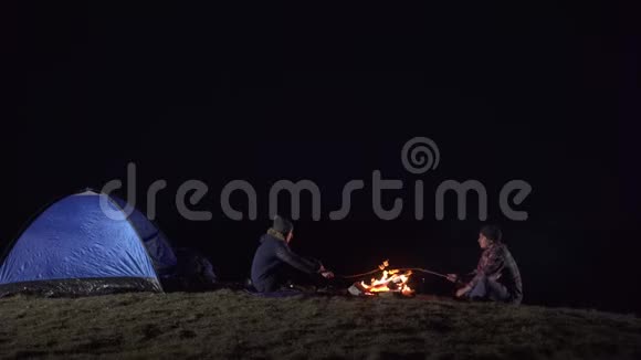 游客们坐在帐篷附近晚上在山上的火堆上煎棉花糖视频的预览图