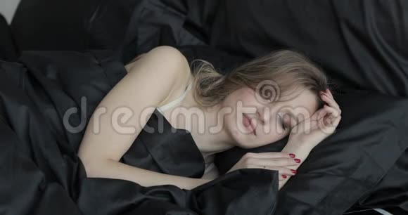 睡在黑色枕头和黑色毯子下的迷人的金发年轻女人的肖像她在说话视频的预览图
