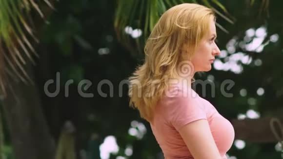 一个美丽的金发女人在一个刮风的日子里站在棕榈树上她看着镜头微笑视频的预览图