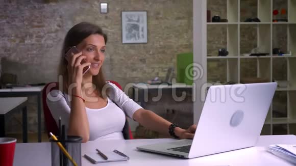 漂亮快乐的女办公室职员在电话里笑着在工作场所的笔记本电脑上打字视频的预览图