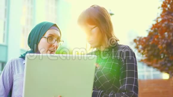 穆斯林女学生戴着头巾她的朋友带着笔记本电脑在公园聊天阳光明媚慢节奏视频的预览图