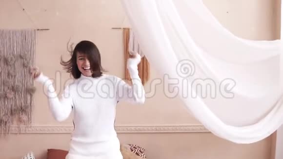 可爱的年轻漂亮女孩穿着白色衬衫和跳线在床上跳跃慢动作视频的预览图