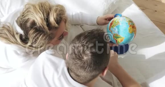 一对恋爱中的情侣在床上看着地球旋转着它指着随机选择一个蜜月旅行的地方视频的预览图