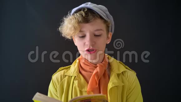 年轻时尚的女人在贝雷帽与卷曲的金发阅读书隔离在黑色背景女性穿黄色夹克视频的预览图
