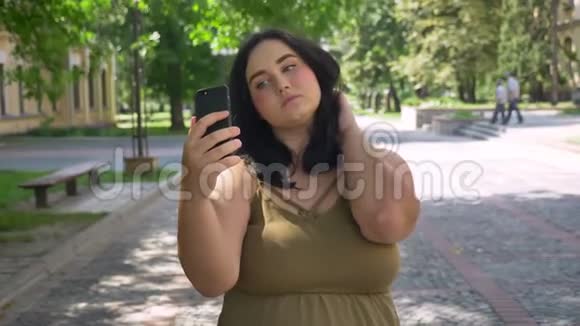 美丽的年轻超重女士用手机自拍并在镜头中做不同的脸站在街上视频的预览图