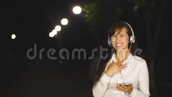 夜晚女孩在城市公园里走着灯笼灯戴着耳机听音乐的漂亮女孩视频的预览图