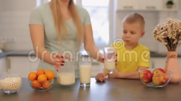 美丽的白种人妈妈留着长长的白发为她穿着黄色衬衫的可爱儿子倒了一杯牛奶现代视频的预览图