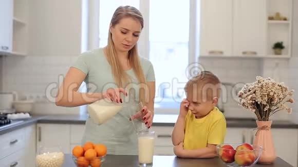 美丽的白种人妈妈留着长长的白发为她穿着黄色衬衫的可爱儿子倒了一杯牛奶现代视频的预览图