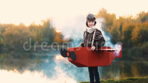 快乐的小飞行员女孩站在日落湖穿着纸板飞机服装彩烟玩飞行员慢动作视频的预览图