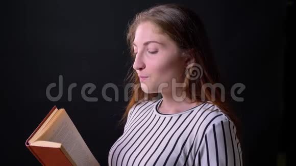 年轻迷人的女性阅读书籍和微笑的相机孤立在黑色工作室背景视频的预览图