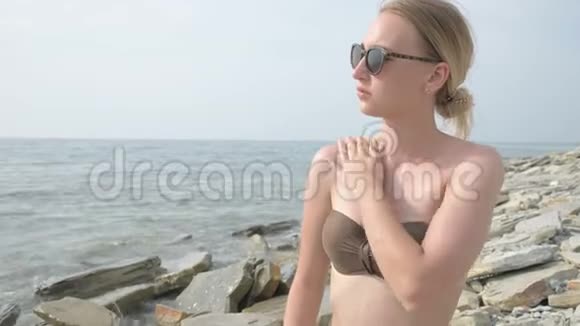 阳光明媚的夏天一个戴太阳镜脖子上涂防晒霜坐在海滨的迷人少女视频的预览图