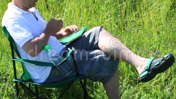 一个男人坐在一个有灯光的草地上的野餐椅上他休息用智能手机工作喝水草和花从年代摇曳而出视频的预览图