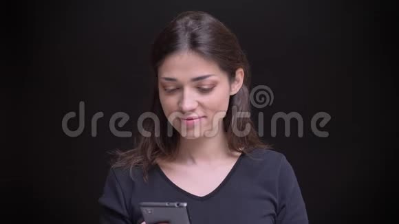 年轻的白种人长发黑发女孩微笑着观看黑色背景的智能手机的肖像视频的预览图