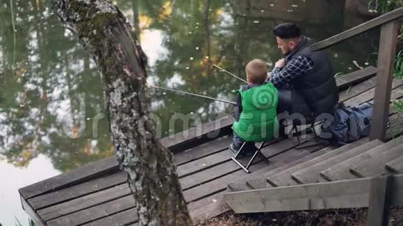 人们父子在森林里的池塘里钓鱼坐在木墩上的椅子上手持木棍视频的预览图