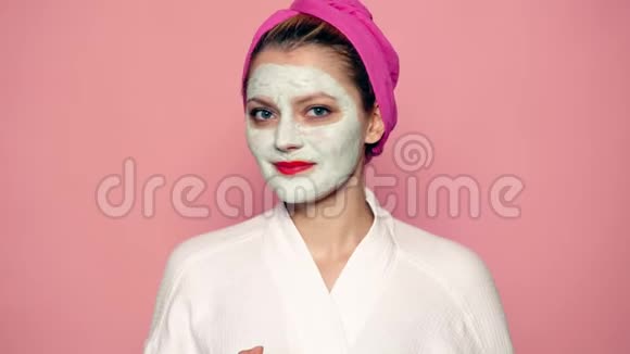 女孩脸上戴着面具头上戴着毛巾在粉红色的背景上扔出一个苹果漂亮的女孩化妆视频的预览图