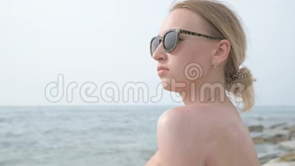 一个年轻迷人的女孩戴着太阳镜脖子和肩膀上涂着防晒霜坐在海边视频的预览图