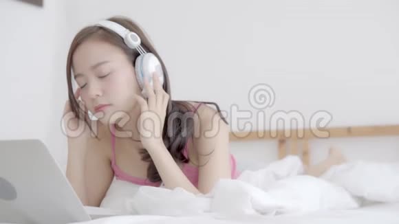 美丽的年轻亚洲女人躺在卧室里用手提电脑放松听音乐女孩在家里看视频聊天视频的预览图