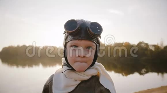 小飞行员男孩穿着复古飞行员服装戴着围巾和眼镜看着相机微笑着看日落湖慢动作视频的预览图