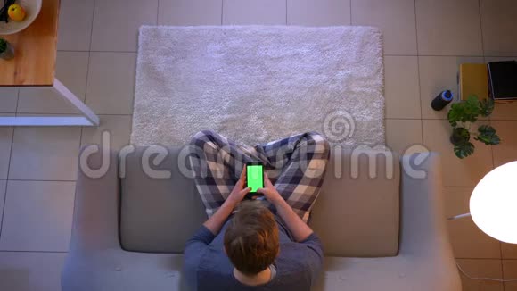 年轻的穿着随意的男性坐在沙发上用带有绿色屏幕的手机进行特写镜头视频的预览图