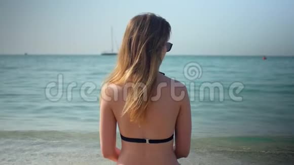 穿比基尼的苗条优雅的女孩正坐在海滩上在阳光明媚的日子里看着大海后景视频的预览图